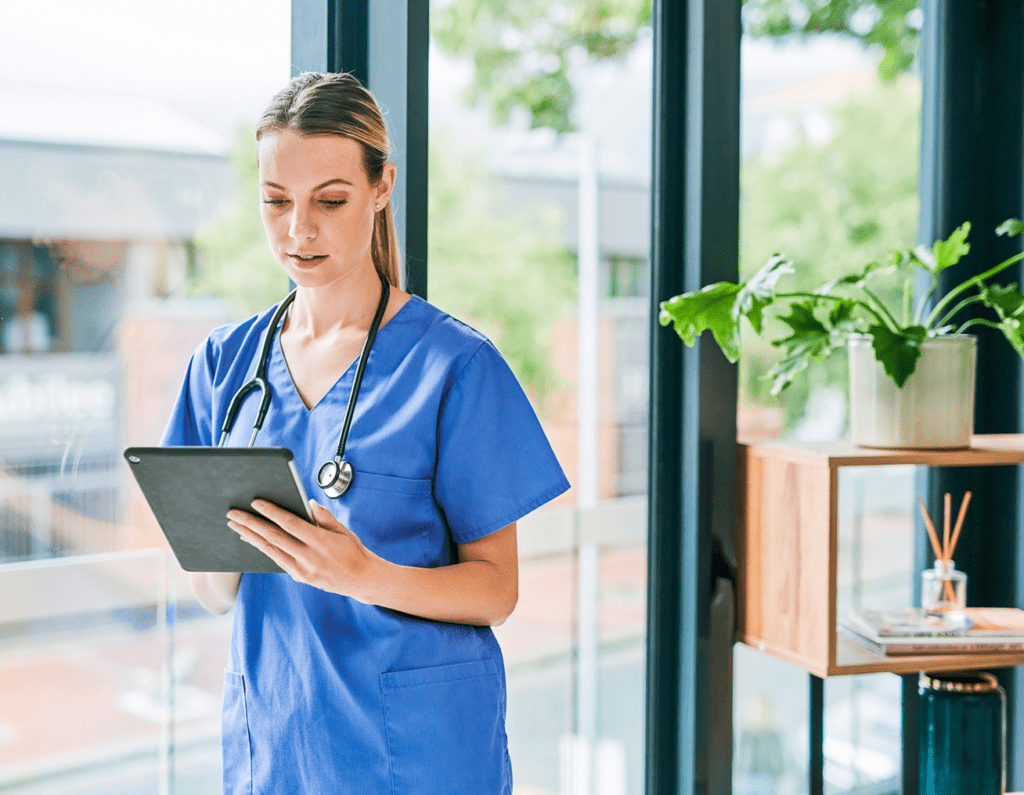 A nurse in blue scrubs reviewing a tablet in a bright room.