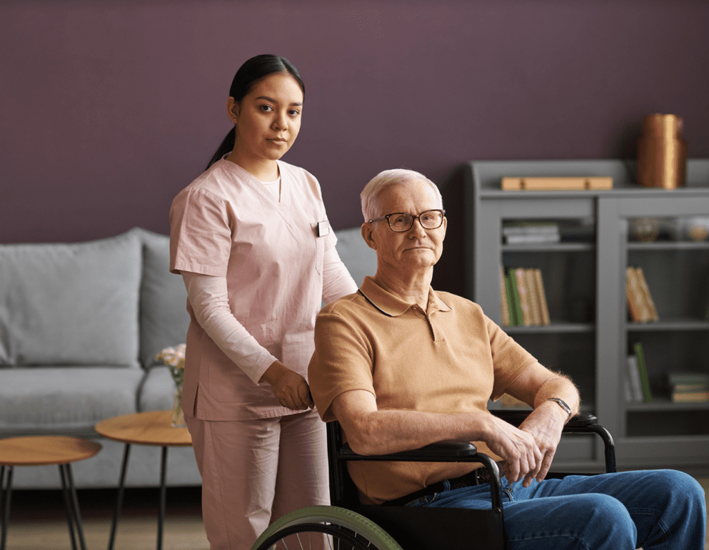 A caregiver stands behind an elderly man in a wheelchair, offering support.
